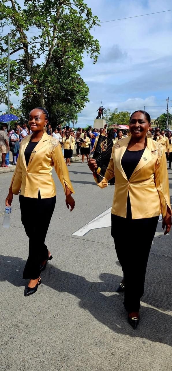 Dos mujeres con sacos dorados de satín y pantalón negro desfilando como parte de un grupo uniformado.