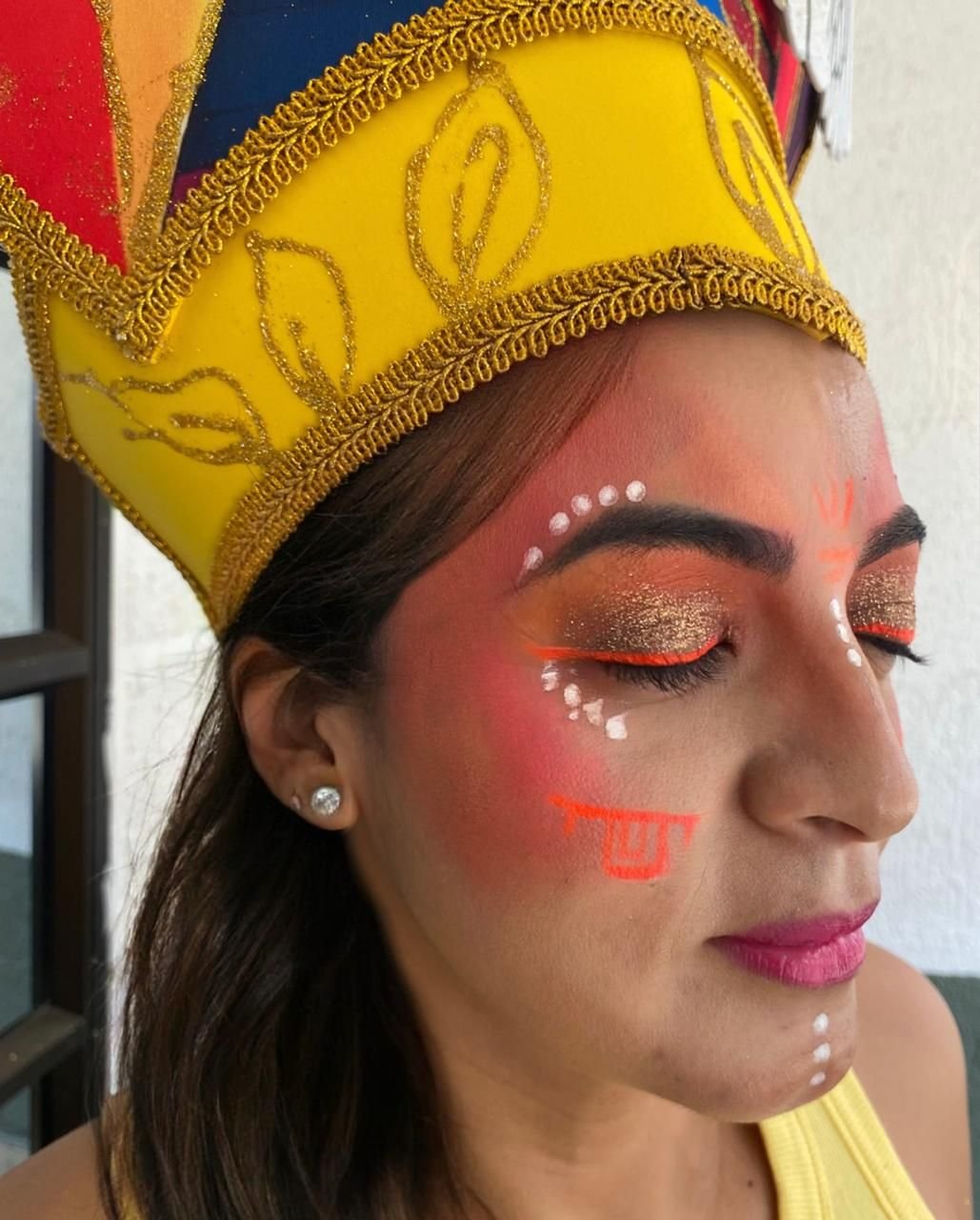 Mujer de perfil con corona amarilla de bordes dorados y maquillaje artístico en tonos naranja y blanco.