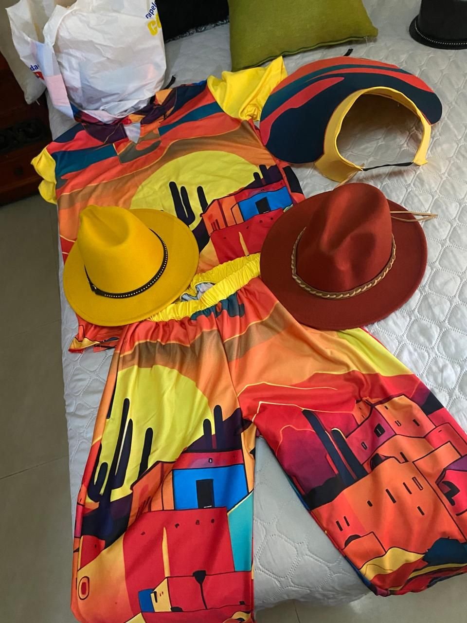 Conjunto de blusa y pantalón con estampado de atardecer y casas de colores, junto a dos sombreros.