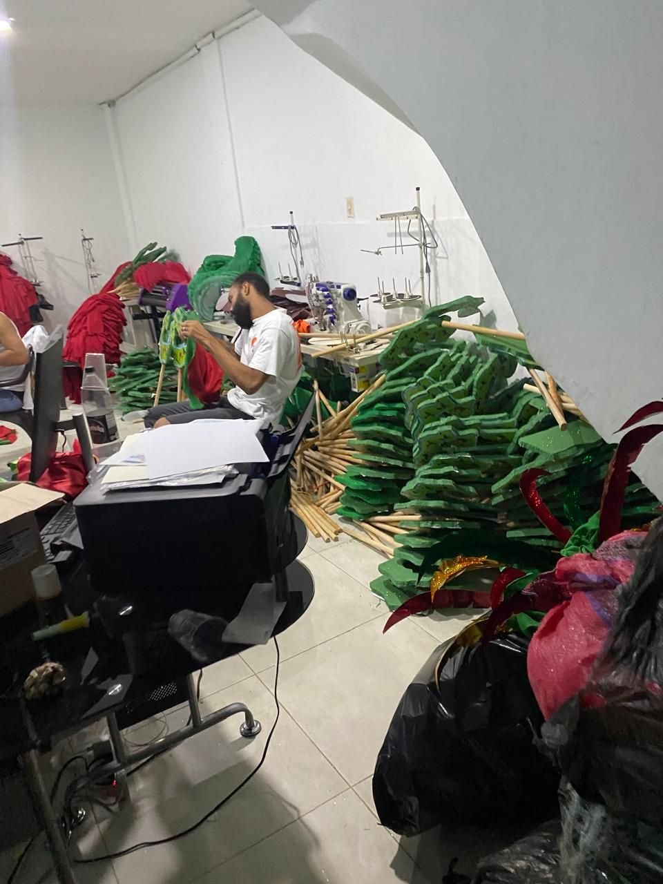 Hombre cosiendo en el taller junto a montones de piezas verdes de foami y telas rojas.