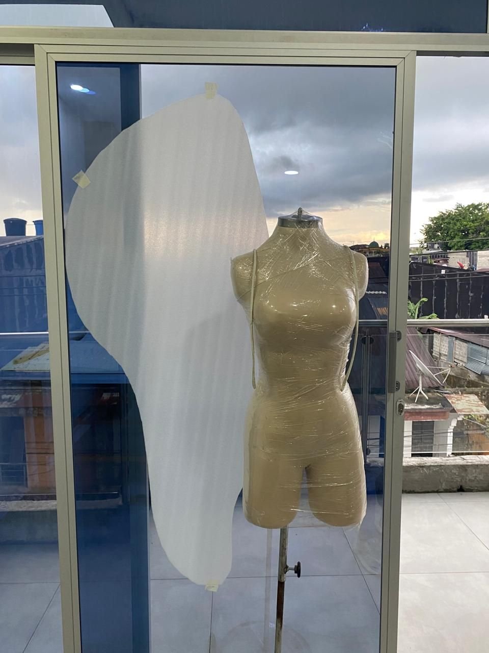 Maniquí cubierto con plástico y una pieza de foami cortada sobre un ventanal.